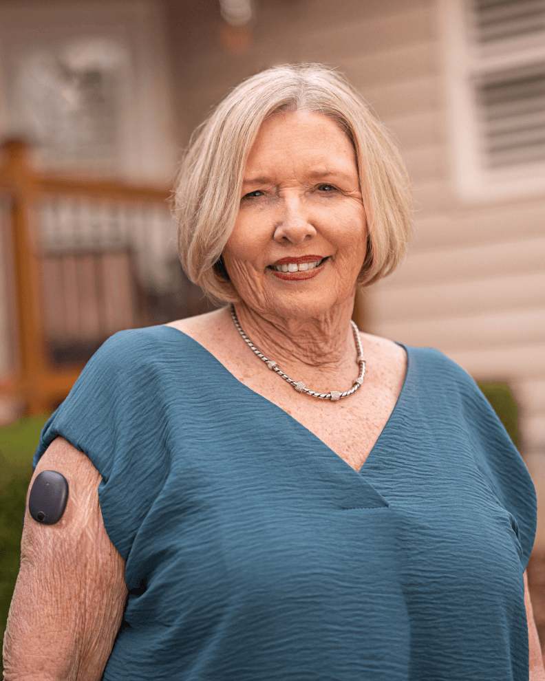 An Eversense user with short blonde hair, smiling, wearing a blue top, and a medical CGM sensor on her upper arm, outside house.